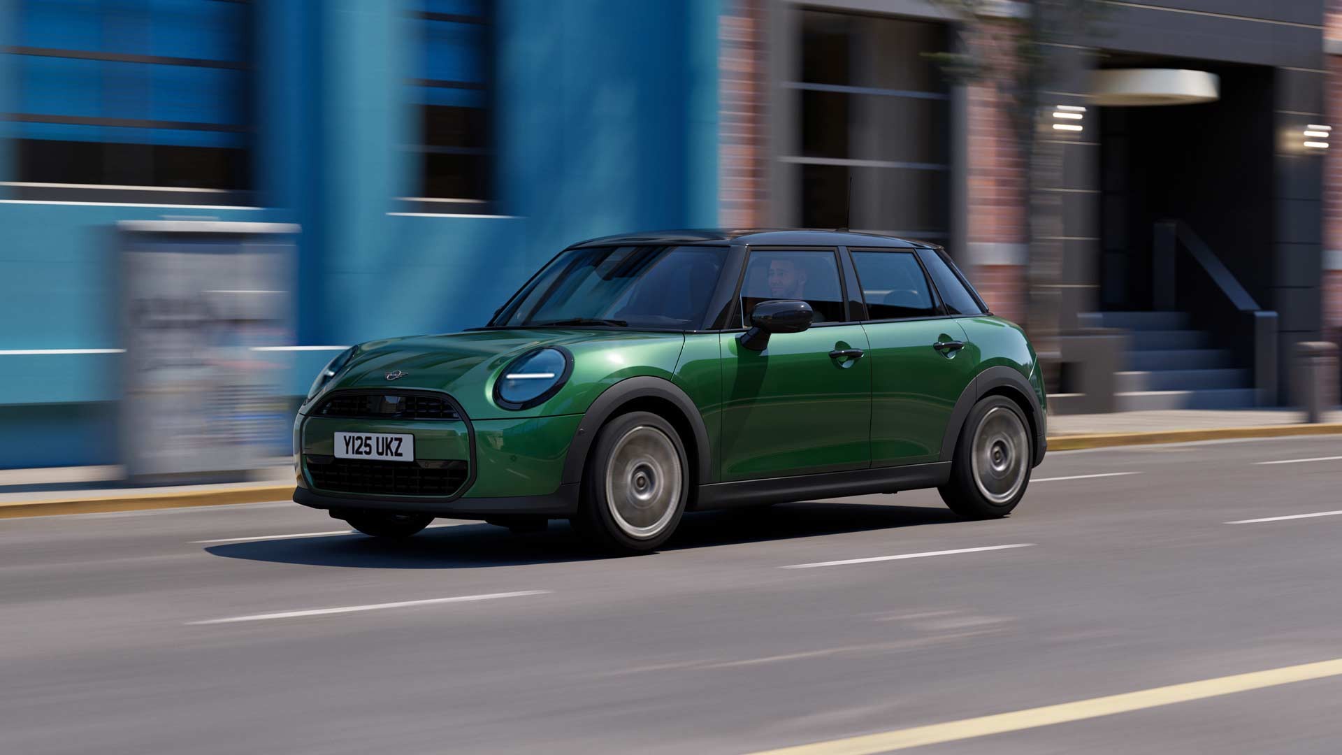 MINI Cooper 5-Door drives swiftly down a city street, passing a bright blue wall and brick building. The scene conveys a sense of speed and urban energy.